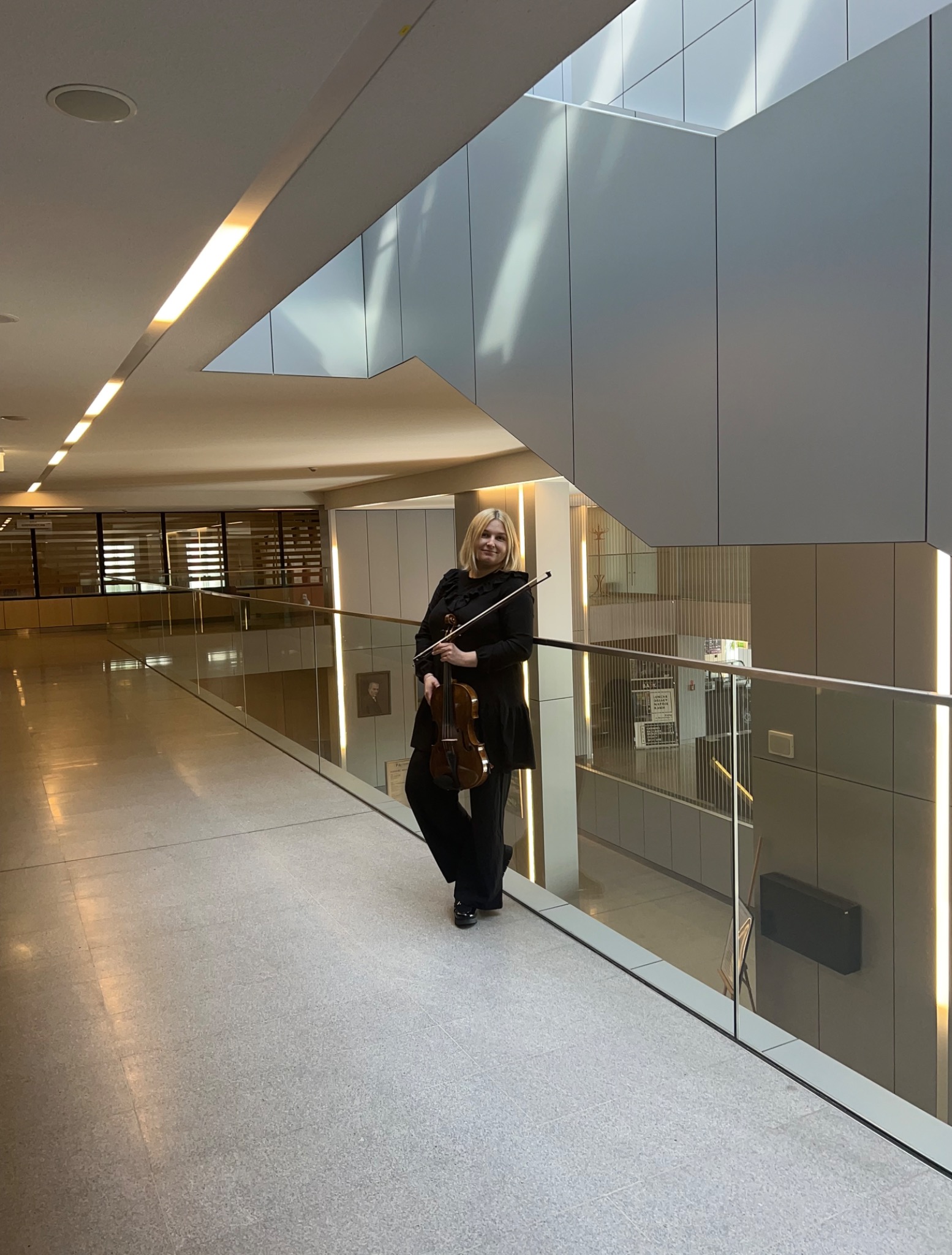 Kobieta z blond włosami w czarnym stroju trzyma skrzypce i smyczek, stojąc na korytarzu z przeszkloną balustradą w nowoczesnym budynku.