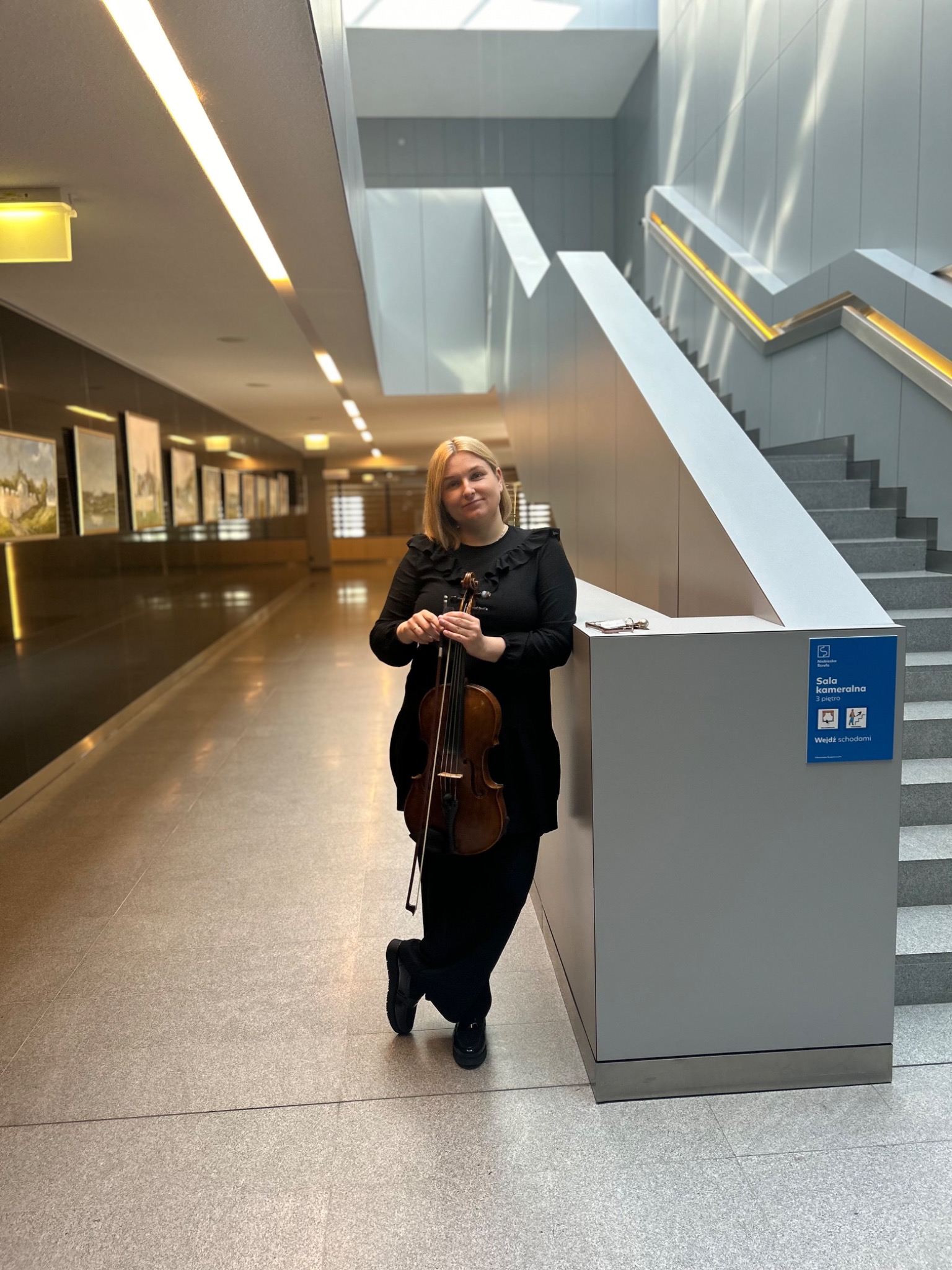 Blondwłosa kobieta w czarnej sukience trzyma skrzypce w korytarzu z obrazami i schodami.
