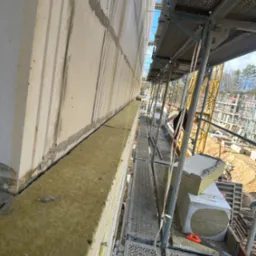 Ocieplanie ściany zewnętrznej budynku z użyciem wełny mineralnej, widok ze rusztowania, żółta nić poziomująca, w tle plac budowy z dźwigiem.