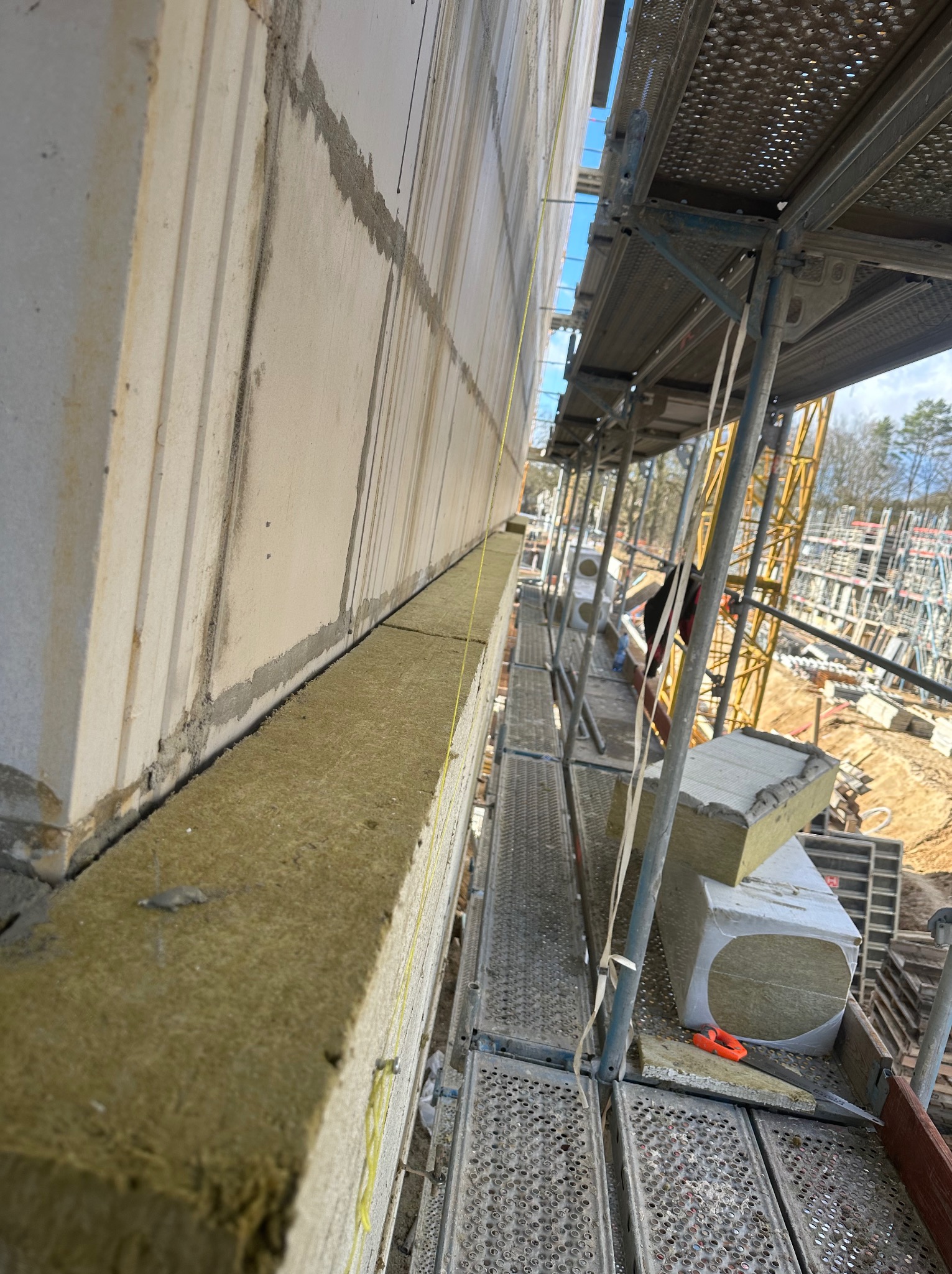 Ocieplanie ściany zewnętrznej budynku z użyciem wełny mineralnej, widok ze rusztowania, żółta nić poziomująca, w tle plac budowy z dźwigiem.