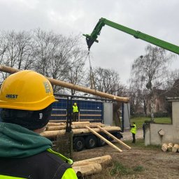 Nest Homes - Montaż domu z bali: robotnicy w kaskach i kamizelkach, dźwig podnosi belkę z ciężarówki. Widok zza pracownika w kasku Nest Homes.