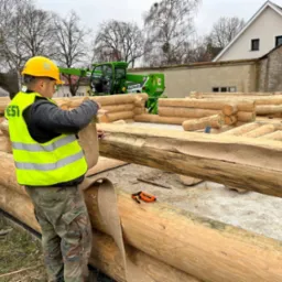 Pracownik w żółtym kasku układa izolację między balami na budowie. W tle widoczny dźwig teleskopowy i budynki.