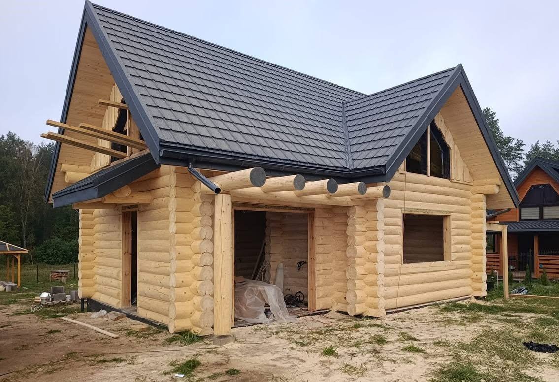 Surowy dom z bali w trakcie budowy, z ciemnoszarą dachówką, widoczne elementy konstrukcyjne i przygotowane otwory okienne, w tle zieleń i drugi dom.