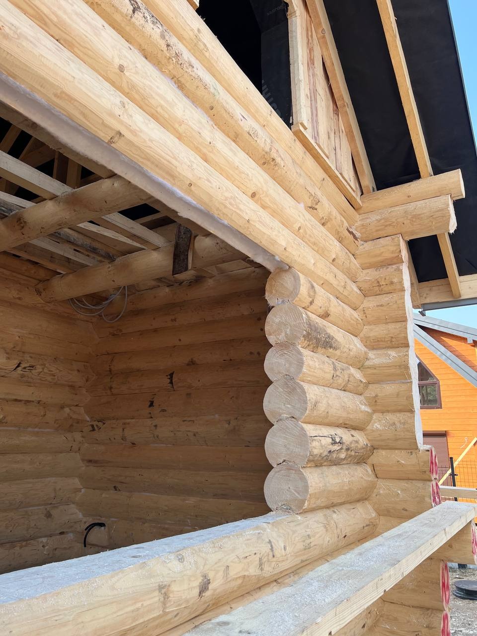 Wnętrze w surowym stanie domu z bali w trakcie budowy, widoczne jasne drewno i konstrukcja dachu z czarną membraną. Częściowo widoczny pomarańczowy budynek w tle.