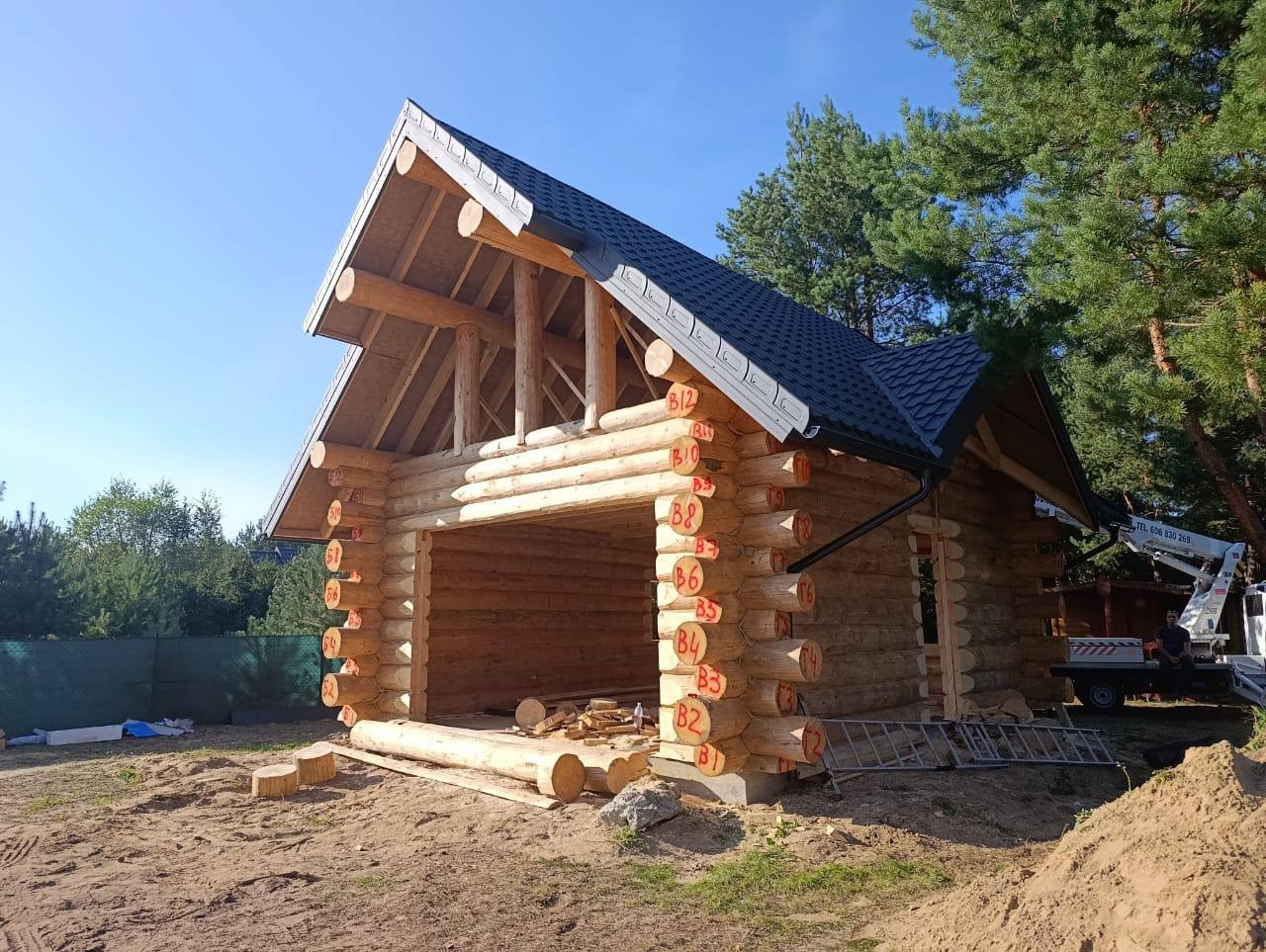 Surowy stan domu z bali z numeracją bali na zewnątrz, z czarnym dachem i widocznym sprzętem na zewnątrz. Niebieskie niebo i drzewa w tle.