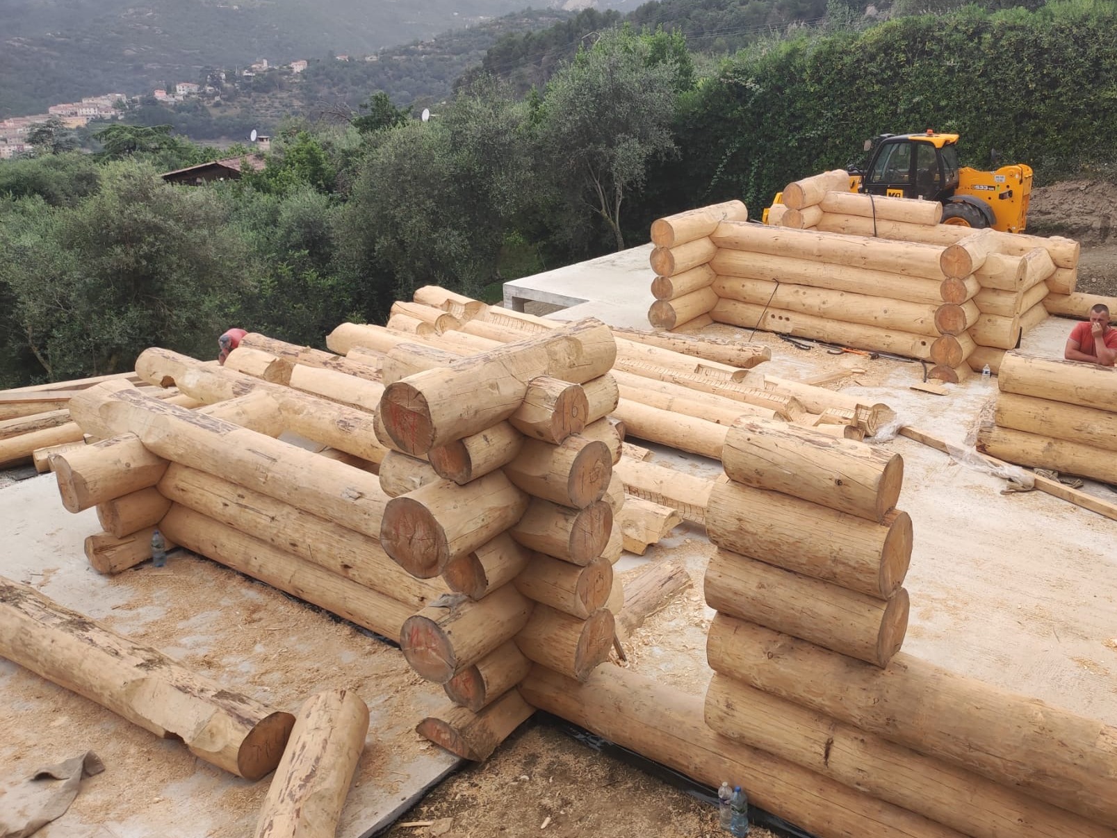 Budowa domu z bali: fragment ściany z okrąglaków, widoczny operator koparki i otoczenie z zielenią. Etap budowy z wykorzystaniem ciężkiego sprzętu.