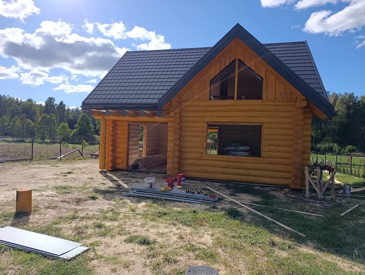 Surowy stan domu z bali z ciemną dachówką, widok z zewnątrz, w tle zieleń drzew i błękitne niebo. Na pierwszym planie narzędzia i materiały budowlane.