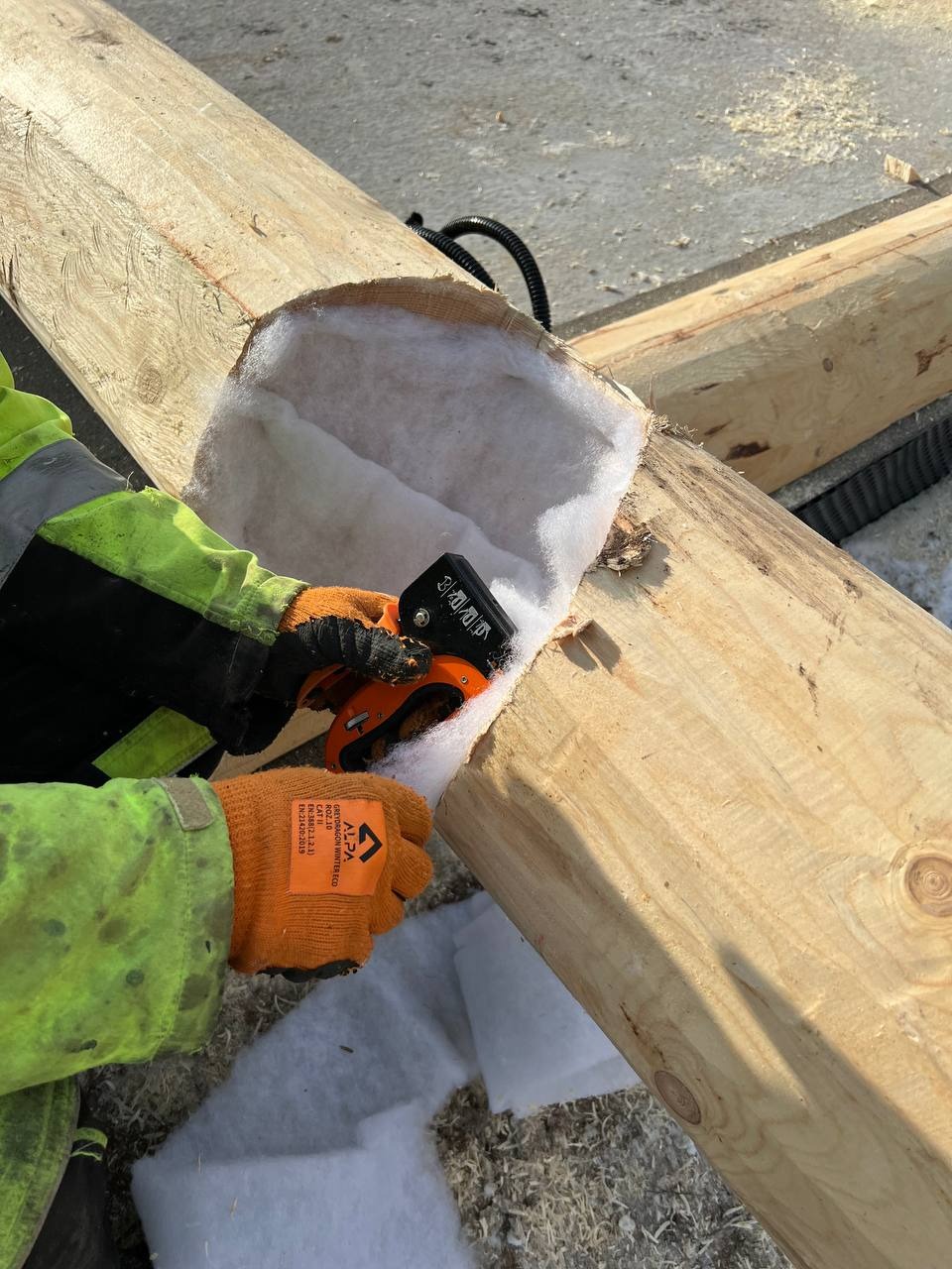 Ocieplanie bala wełną mineralną. Pracownik w rękawicach docina izolację termiczną przy użyciu noża. Widoczne drewniane bale i fragment instalacji.