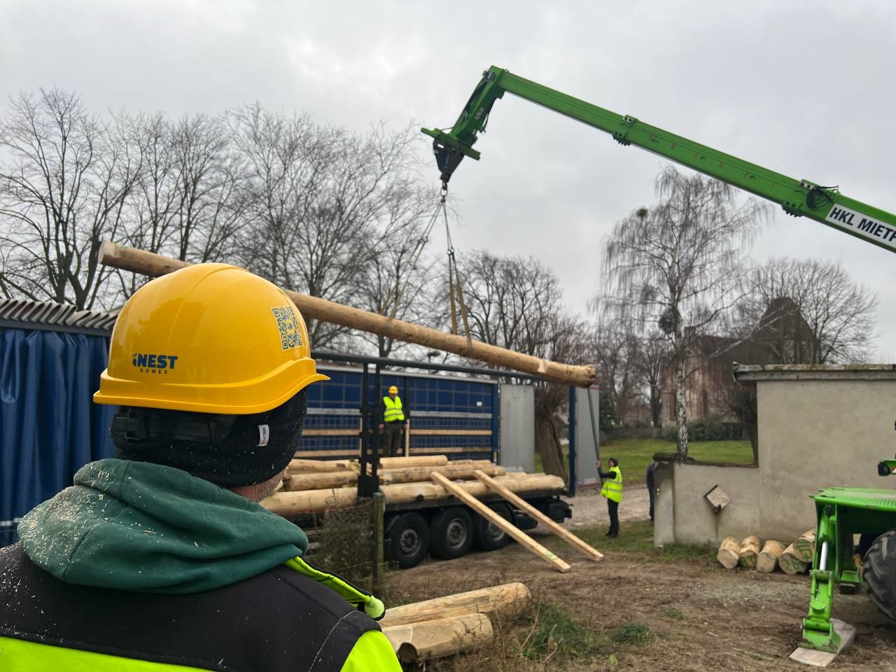 Montaż domu z bali: robotnicy w kaskach i kamizelkach, dźwig podnosi belkę z ciężarówki. Widok zza pracownika w kasku Nest Homes.