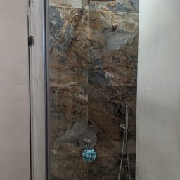 DMT Remonty - Kabina prysznicowa z deszczownicą i baterią, wykończona płytkami imitującymi kamień w odcieniach brązu i szarości, z niebieską myjką zawieszoną na ścianie.