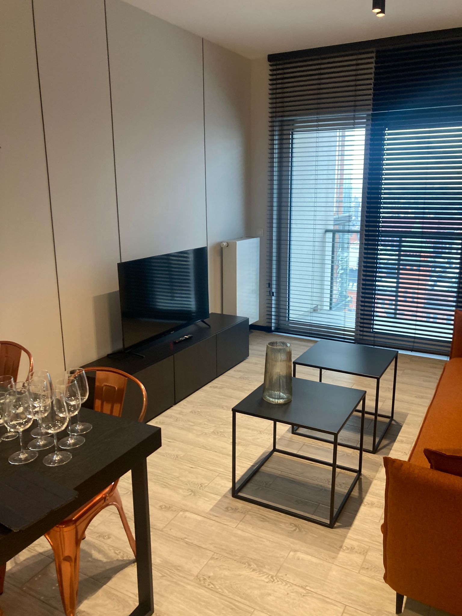 Jasne wnętrze mieszkania z parkietem, minimalistycznym stolikiem kawowym, pomarańczową sofą i stołem z kieliszkami do wina, oświetlone punktowymi lampami sufitowymi i wpadającym światłem przez okno...