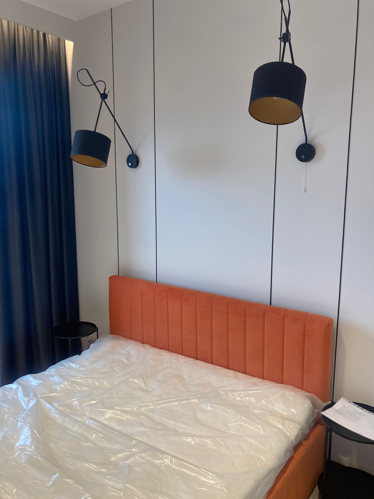 Nowoczesna sypialnia z pomarańczowym zagłówkiem łóżka, czarnymi lampami ściennymi i zasłonami w odcieniach granatu, łóżko przykryte folią ochronną.
