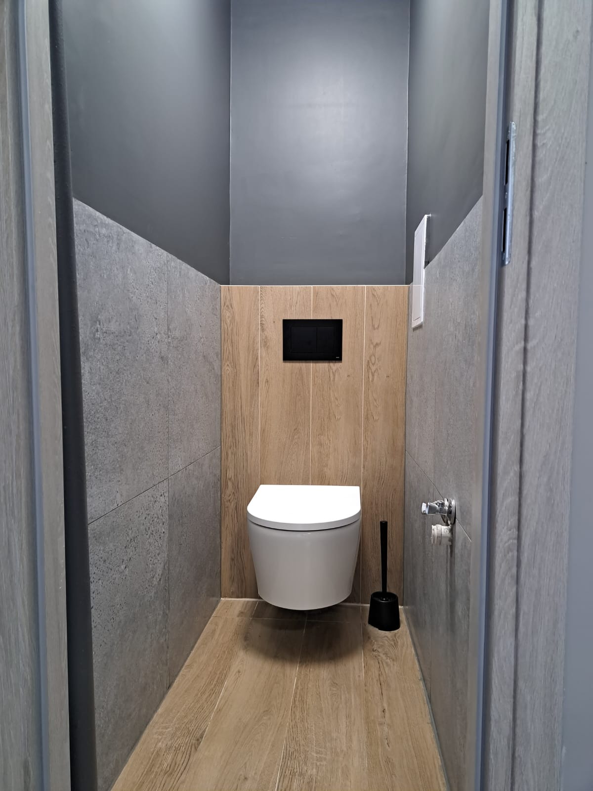 Nowoczesna toaleta podwieszana w łazience wykończonej płytkami imitującymi beton i drewno, z szarą farbą na suficie i czarnym przyciskiem spłuczki.