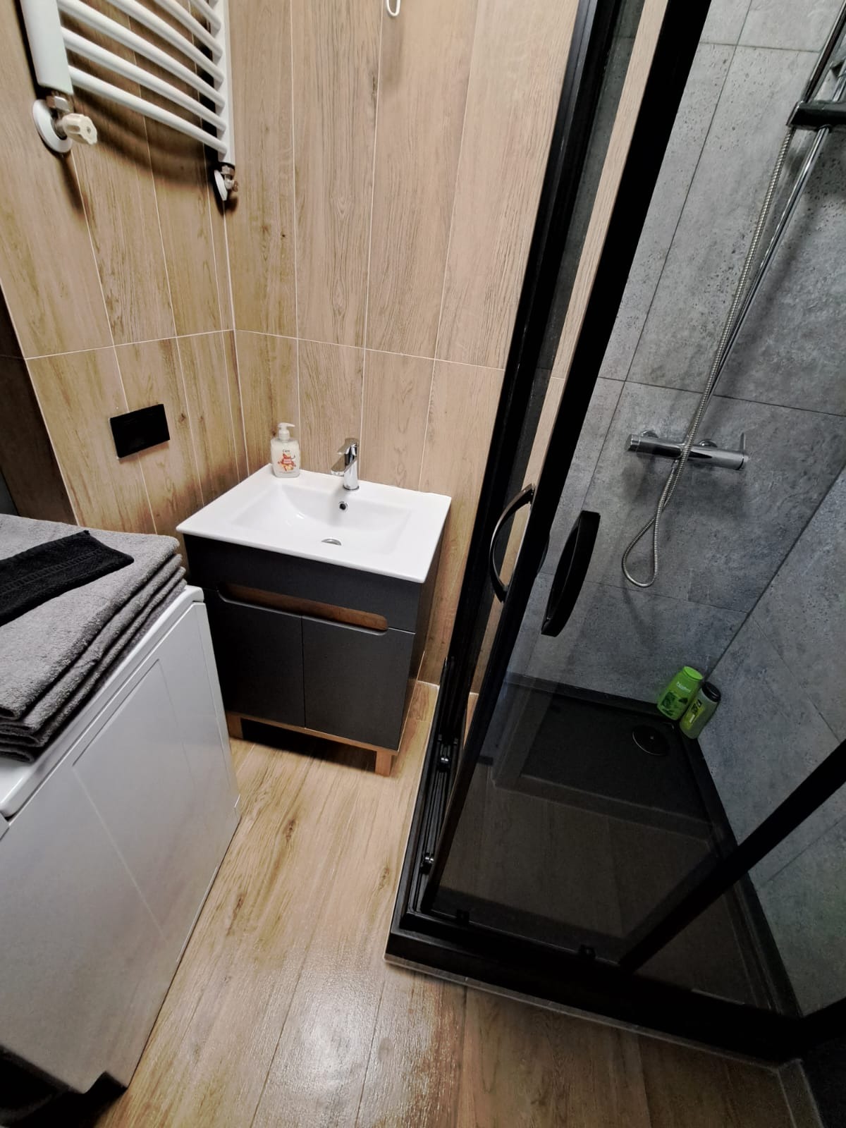 Nowoczesna łazienka z kabiną prysznicową z czarnymi ramami, umywalką z szarą szafką, grzejnikiem drabinkowym i pralką z ułożonymi ręcznikami.