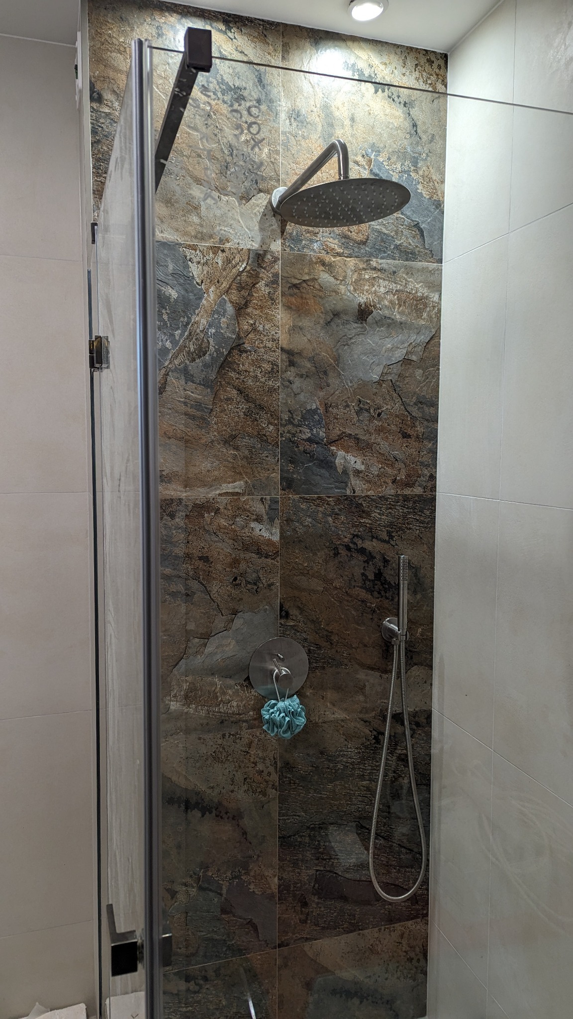 Kabina prysznicowa z deszczownicą i baterią, wykończona płytkami imitującymi kamień w odcieniach brązu i szarości, z niebieską myjką zawieszoną na ścianie.