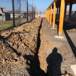 Usługi transportowe i koparka - Wąski wykop pod instalacje biegnący wzdłuż drewnianej konstrukcji z jasnymi słupami, widoczny cień osoby robiącej zdjęcie, w tle siatka zabezpieczająca i budynki.