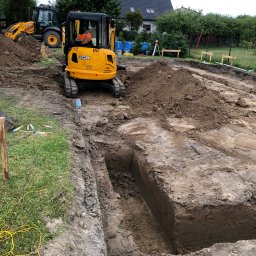 Usługi transportowe i koparka - Żółta mini koparka JCB w trakcie pracy nad wykopem o równych ścianach, w tle widoczna większa koparka i teren budowy z zielenią.