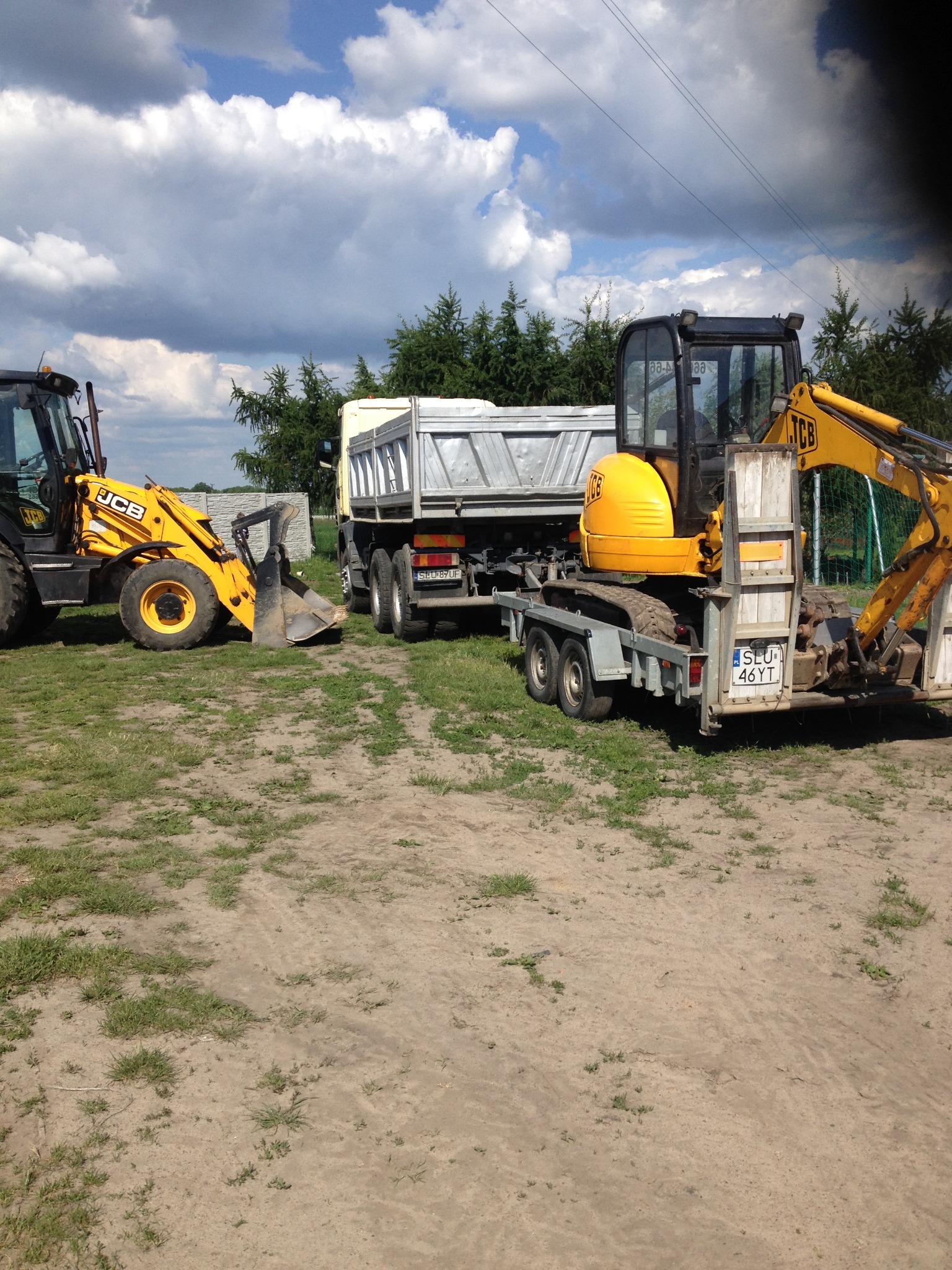 Żółta koparka JCB na przyczepie za ciężarówką, obok druga koparka, na piaszczystym terenie z rzadką trawą i drzewami w tle pod błękitnym niebem z chmurami.