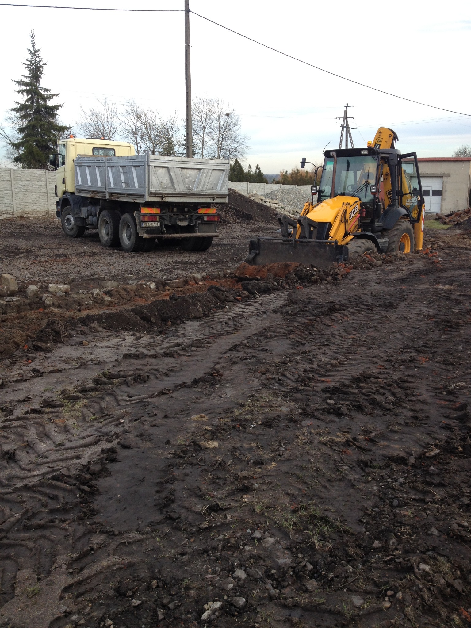 Żółta koparko-ładowarka JCB i wywrotka MAN na placu budowy po deszczu, widoczne ślady opon w błocie, w tle betonowe ogrodzenie i budynek gospodarczy.