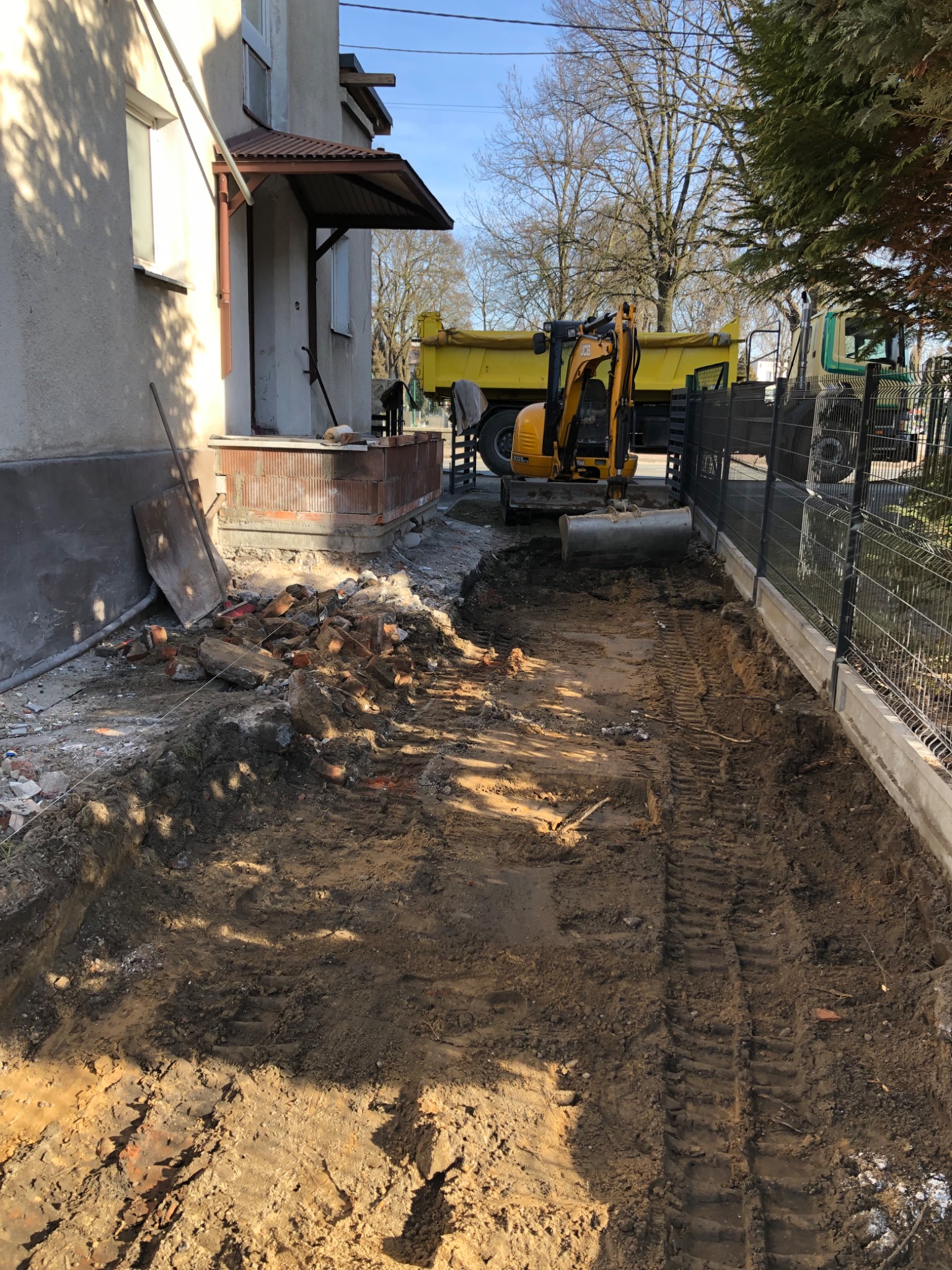 Żółta mini koparka JCB przy wykopie obok budynku, z tyłu wywrotka, widoczne ślady opon na ziemi i fragment fundamentu z cegły.