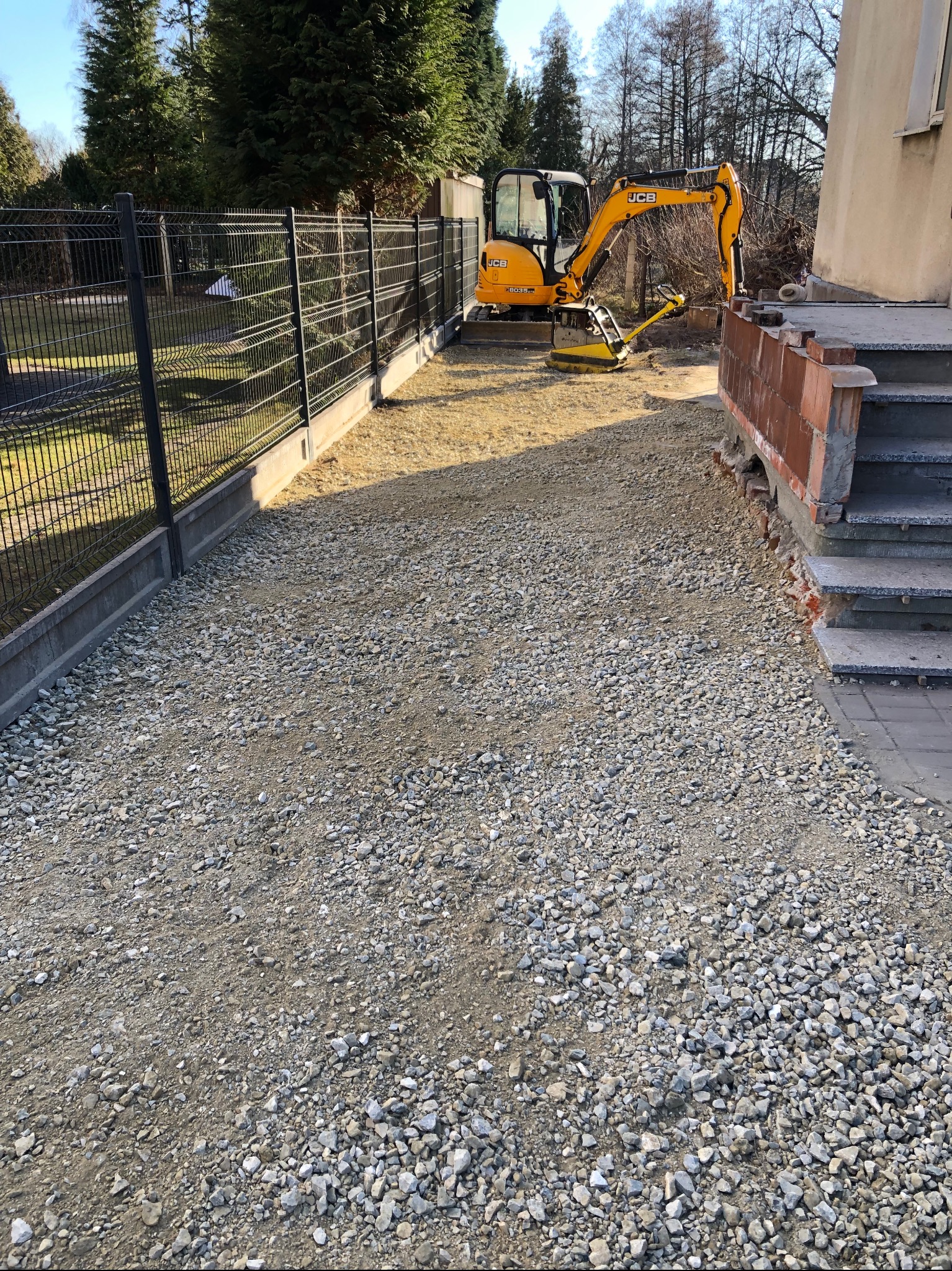 Żółta mini koparka JCB z ubijarką obok schodów budynku i świeżo utwardzonej, szarej, kamienistej powierzchni na posesji z czarnym ogrodzeniem panelowym.
