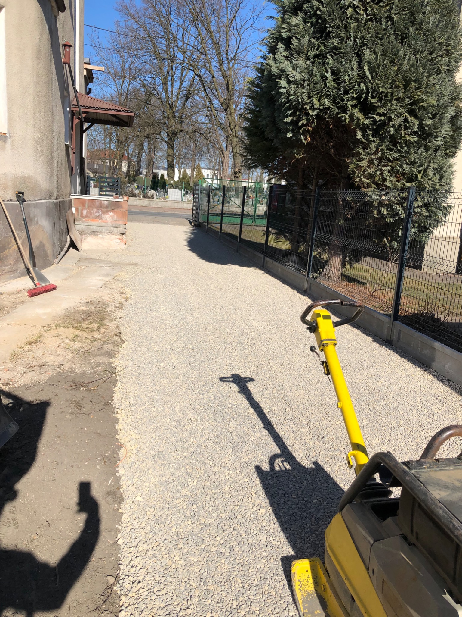 Żółta zagęszczarka wibruje świeżo wysypany żwir na ścieżce, obok budynku i ogrodzenia, z widocznym cieniem operatora.