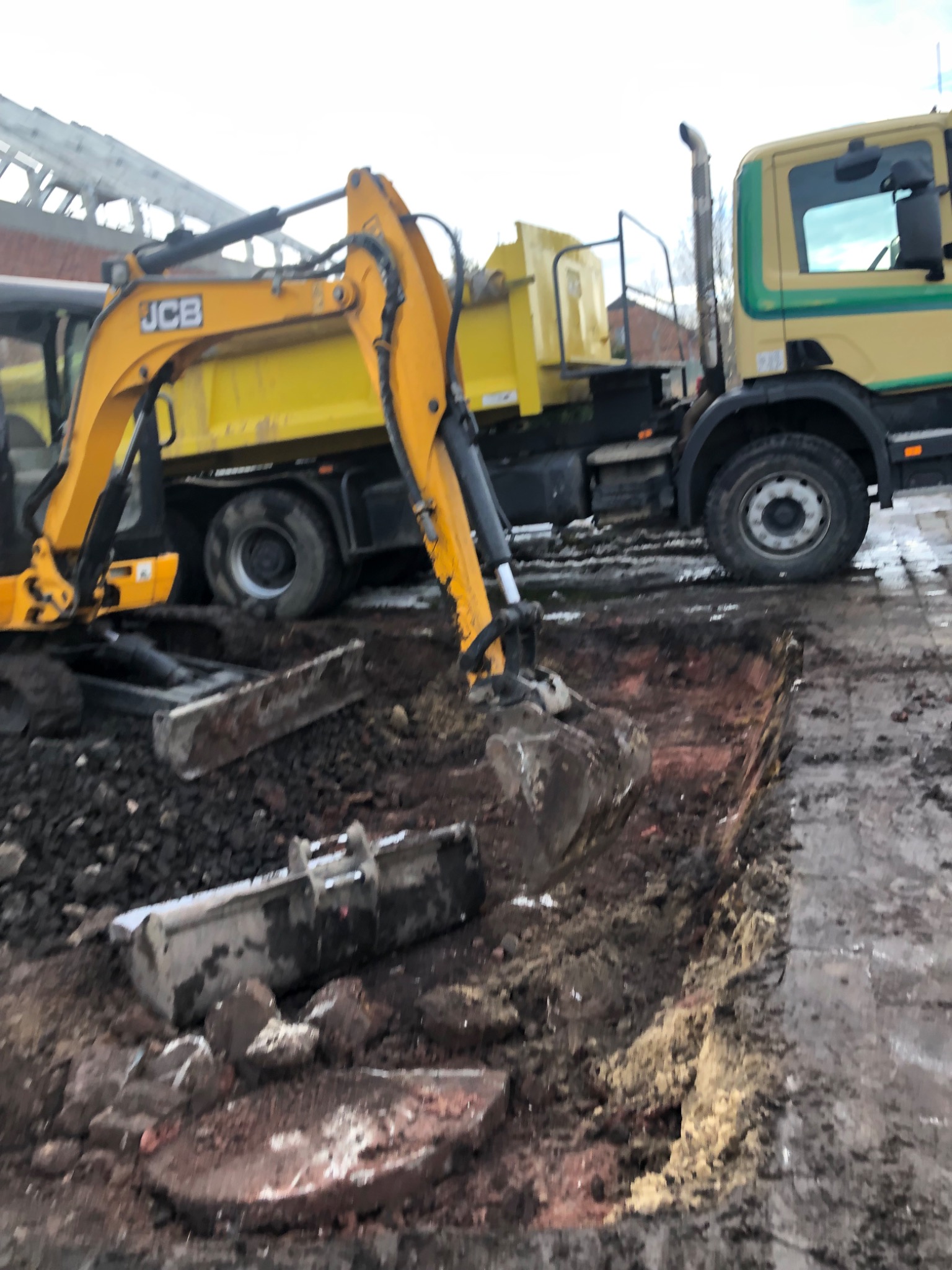 Żółto-czarna koparka JCB w trakcie pracy przy wykopie, obok stoi ciężarówka z żółtą skrzynią ładunkową, teren błotnisty, widoczne ślady opon.