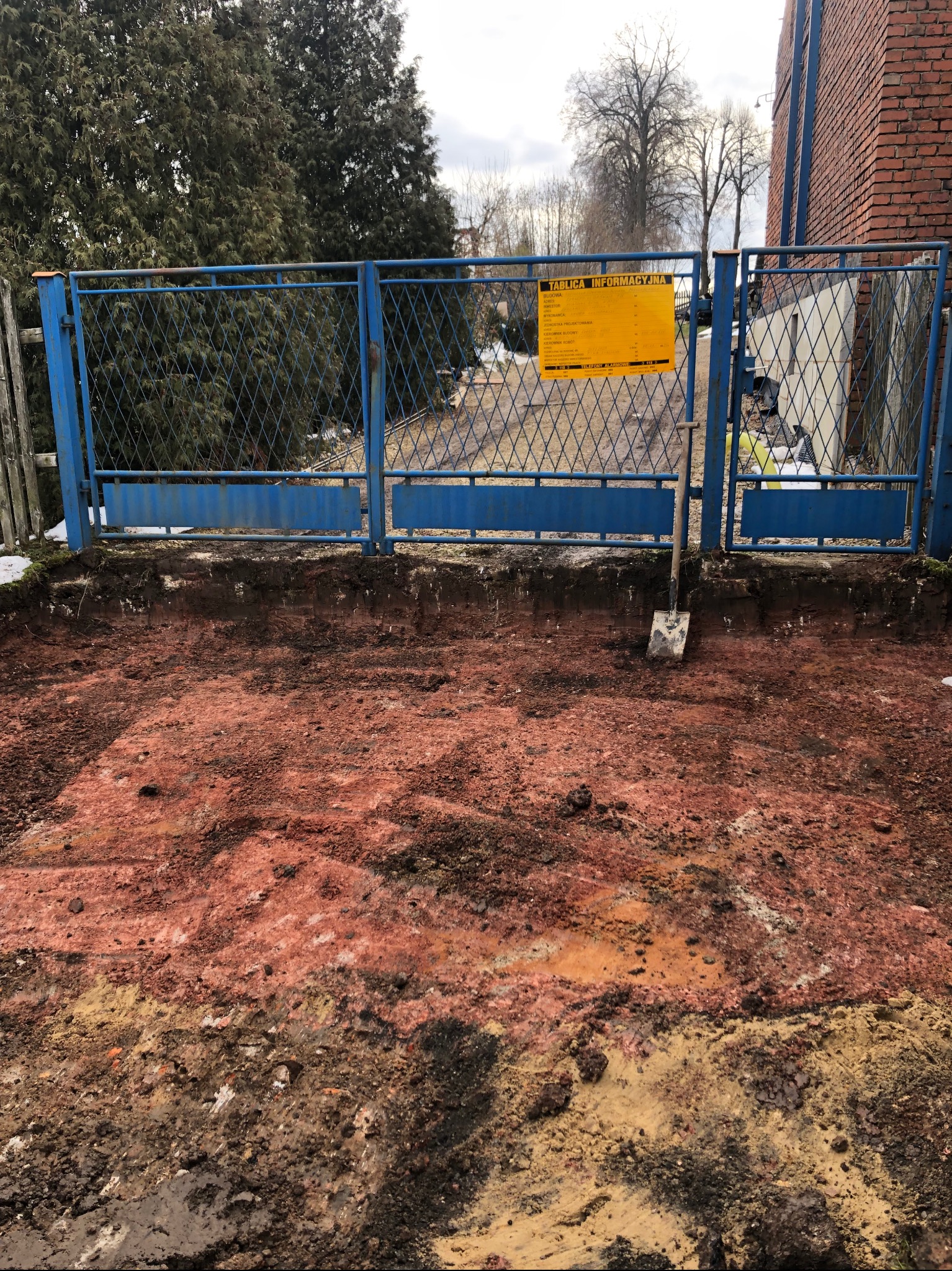Przygotowany teren pod budowę z widoczną warstwą czerwonej ziemi, ogrodzony niebieską siatką i tablicą informacyjną, z łopatą opartą o ogrodzenie.