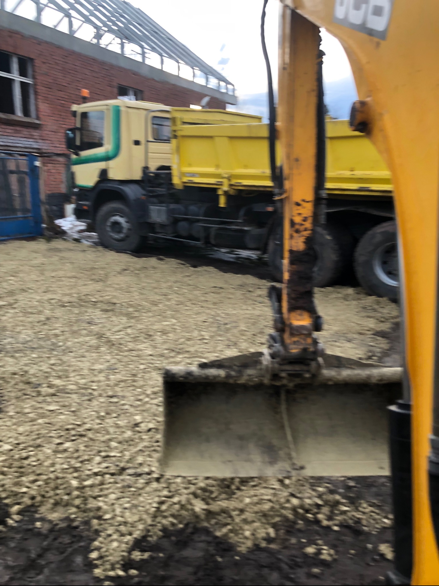 Żółta koparka JCB z łyżką w trakcie pracy, w tle wywrotka i budynek z cegły, teren pokryty drobnym żwirem.