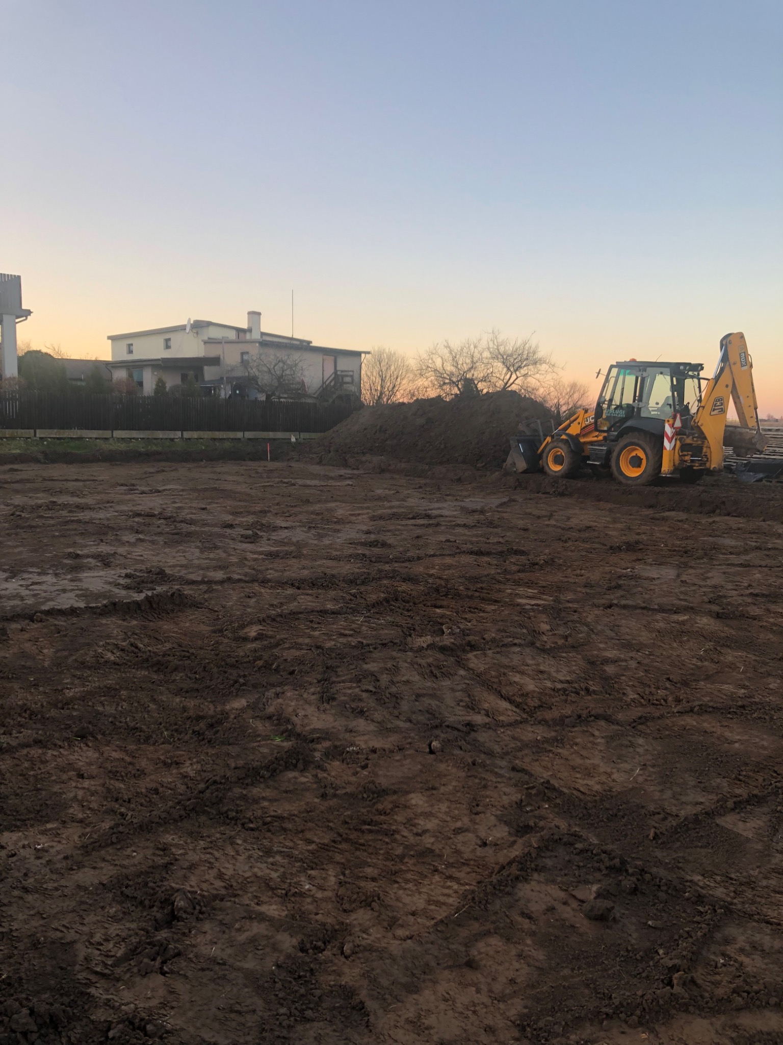 Żółta koparko-ładowarka JCB na wyrównanym terenie w Ciasnej, z pryzmą ziemi w tle i budynkami w oddali, sfotografowana o zmierzchu.