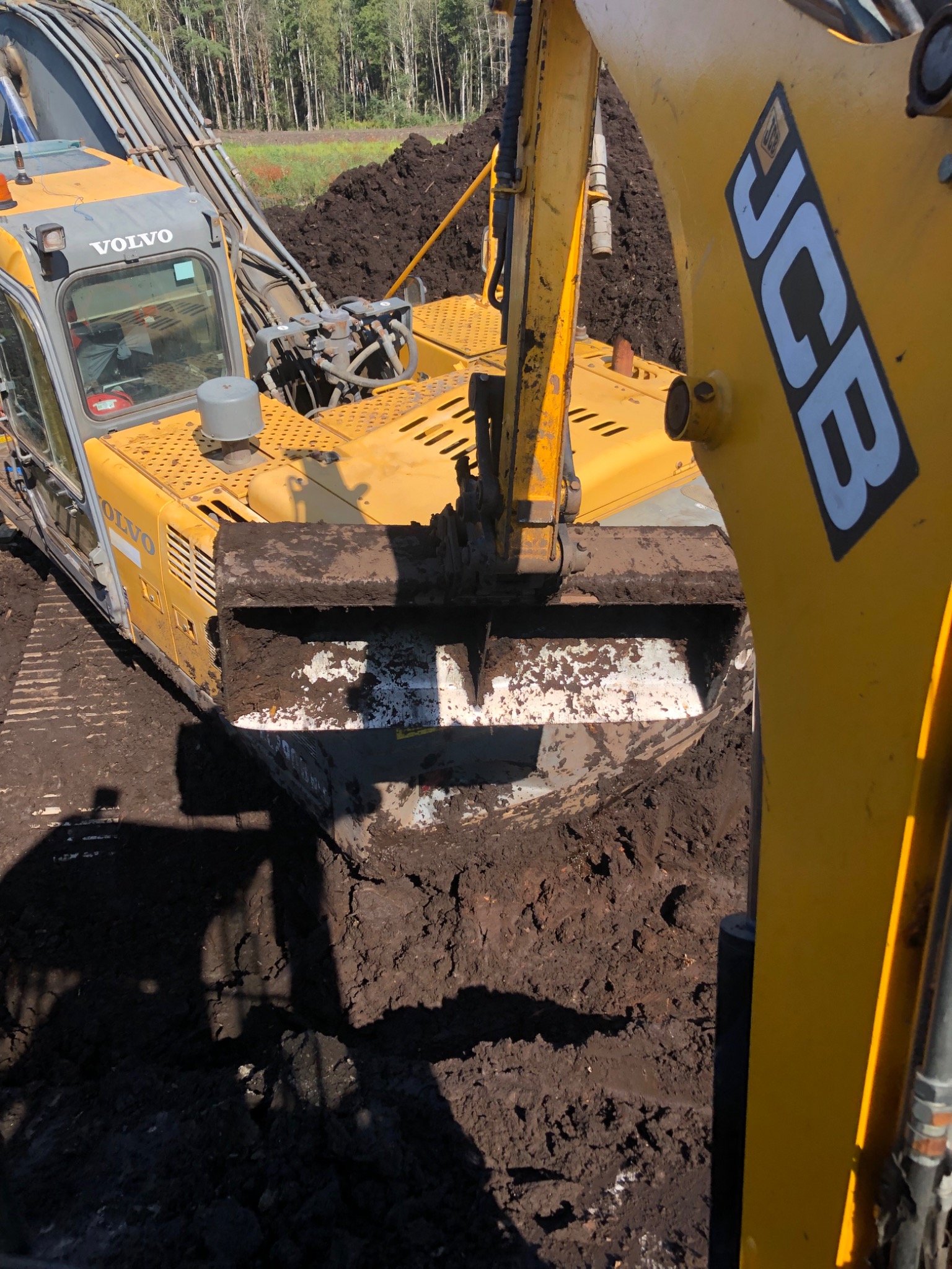 Żółty koparka Volvo i JCB podczas pracy na ziemi, widoczne ślady błota na maszynach.