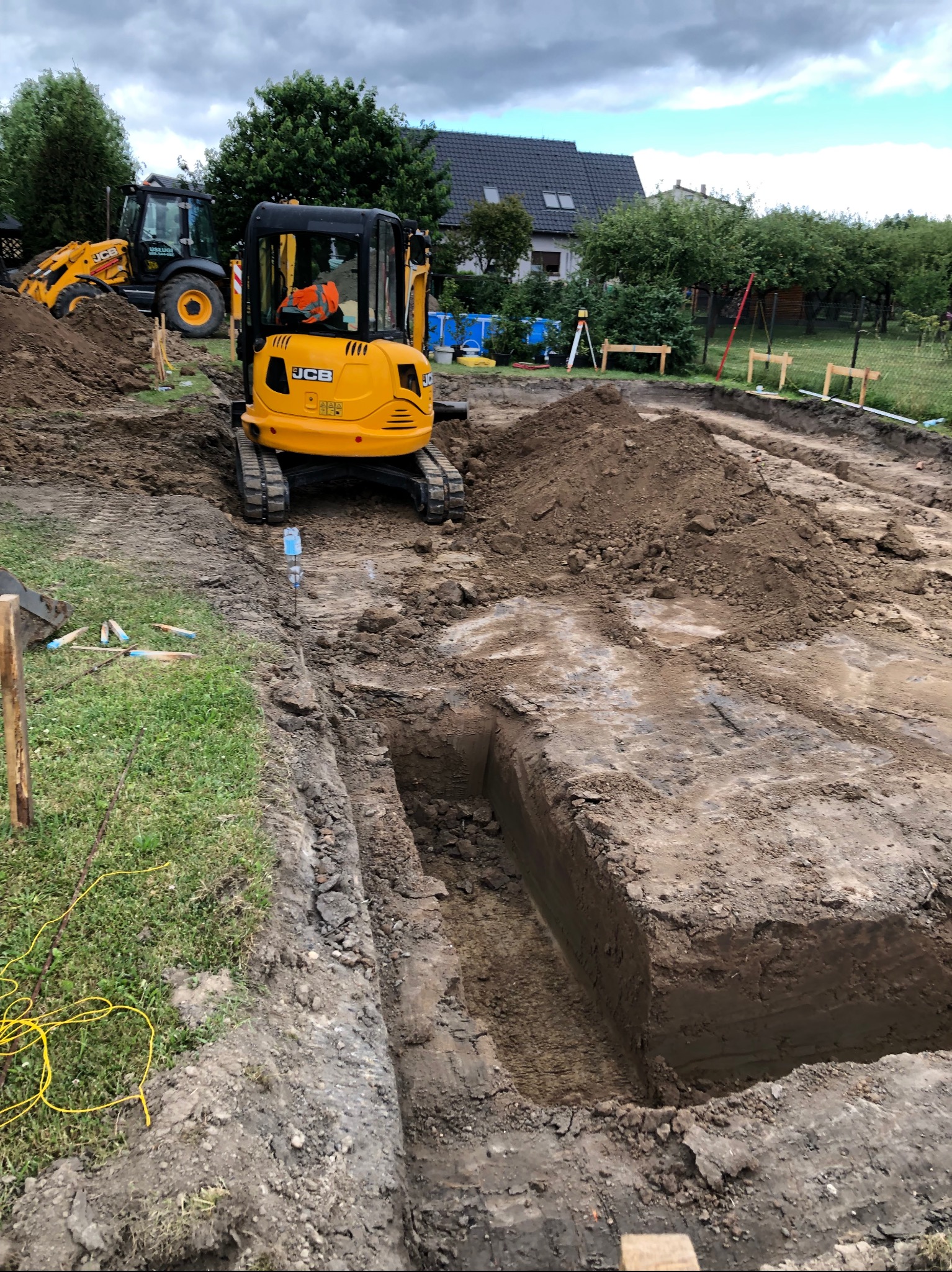 Żółta mini koparka JCB w trakcie pracy nad wykopem o równych ścianach, w tle widoczna większa koparka i teren budowy z zielenią.