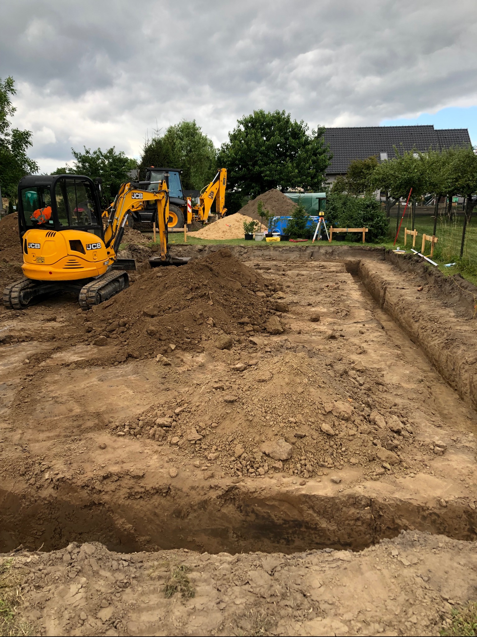 Żółta koparka JCB i wykopane rowy pod fundamenty na placu budowy z widocznym niwelatorem laserowym w tle.
