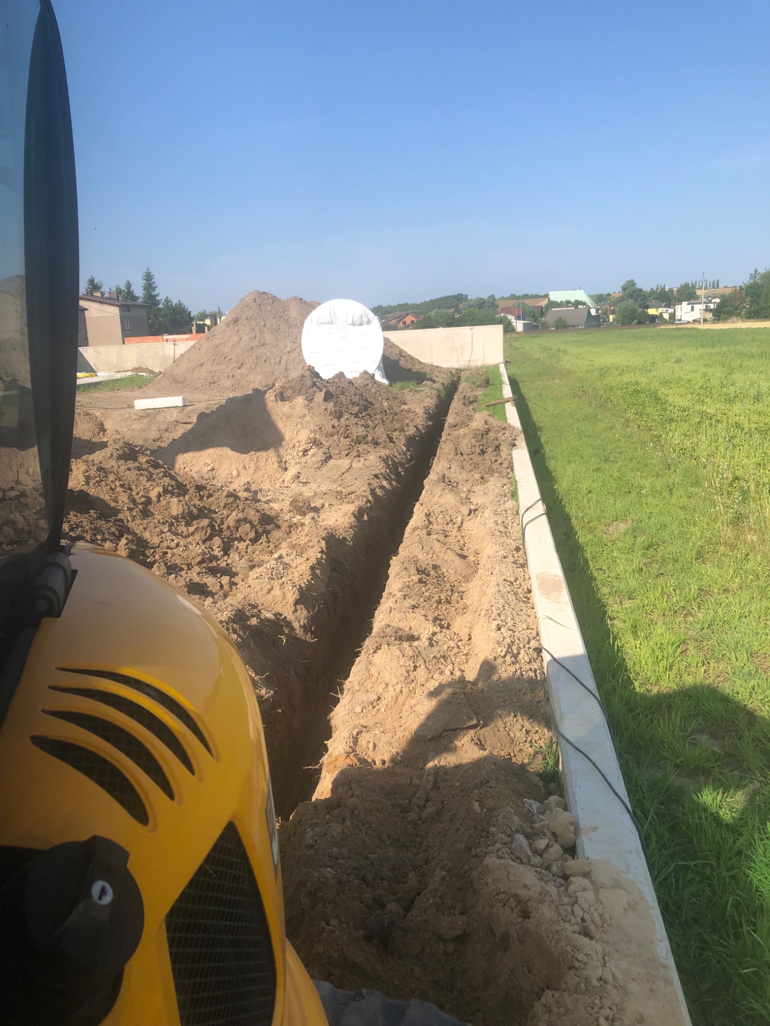 Żółta koparka częściowo widoczna, wykonująca wąski rów w ziemi, prawdopodobnie pod instalacje, z widocznym stosem ziemi i elementami fundamentów w tle.