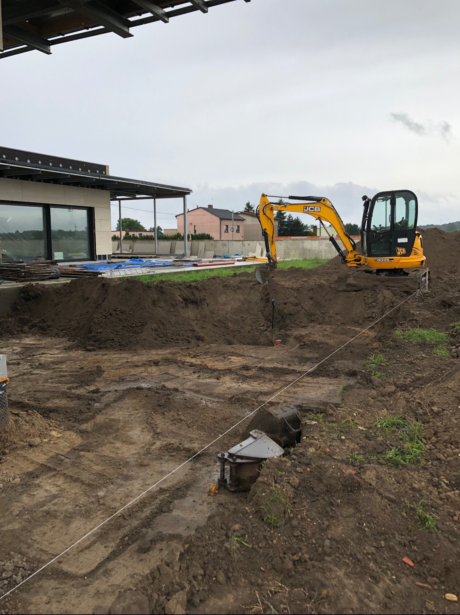 Żółta mini koparka JCB 8035 ZTS wykonuje wykop pod fundament domu jednorodzinnego, widoczny sznurek wyznaczający granice wykopu, w tle budynek w stanie surowym otwartym i pochmurne niebo.