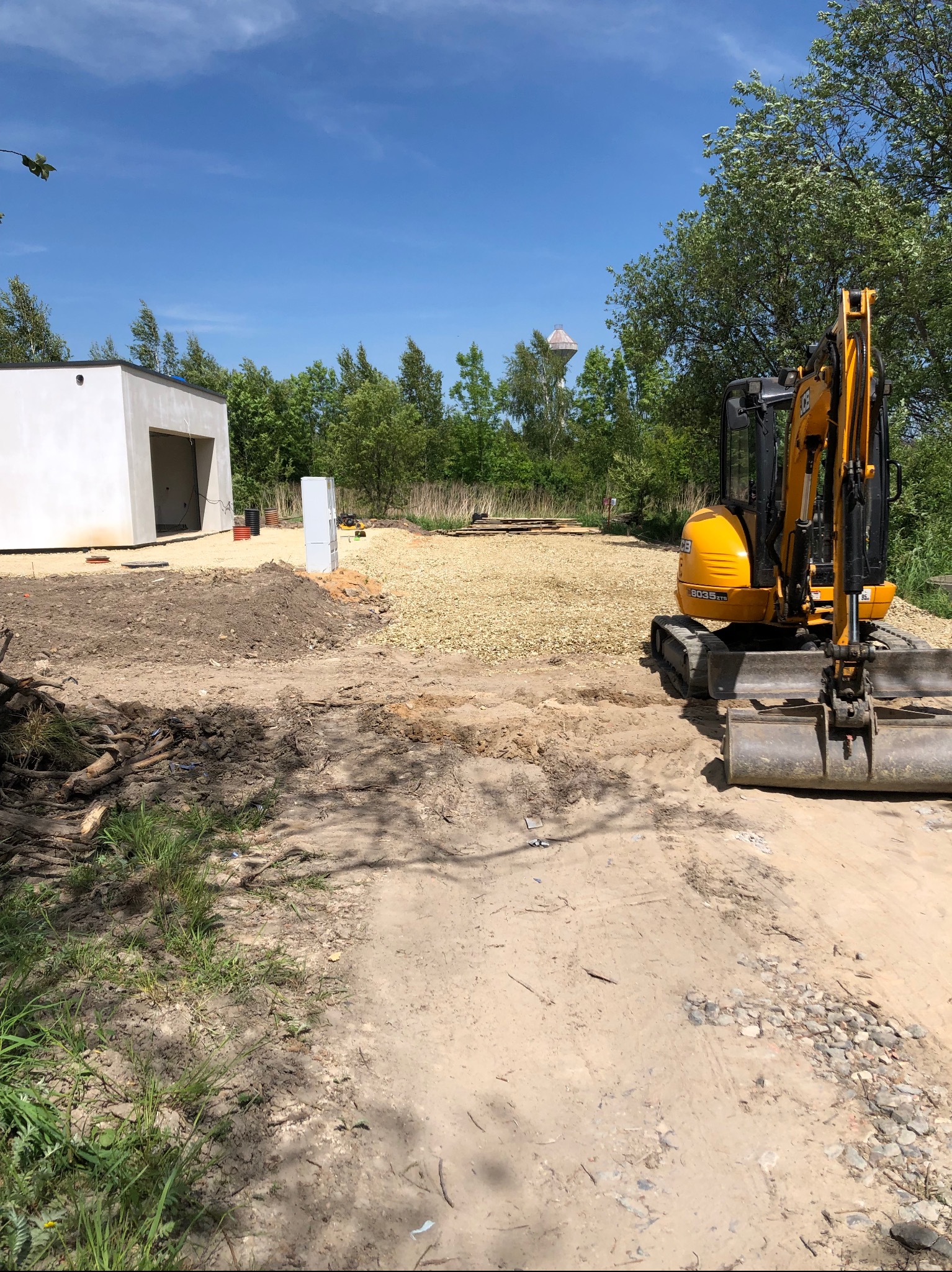 Żółta mini koparka JCB na terenie budowy z częściowo utwardzoną nawierzchnią żwirową, w tle biały budynek w stanie surowym otwartym i wieża ciśnień na horyzoncie.