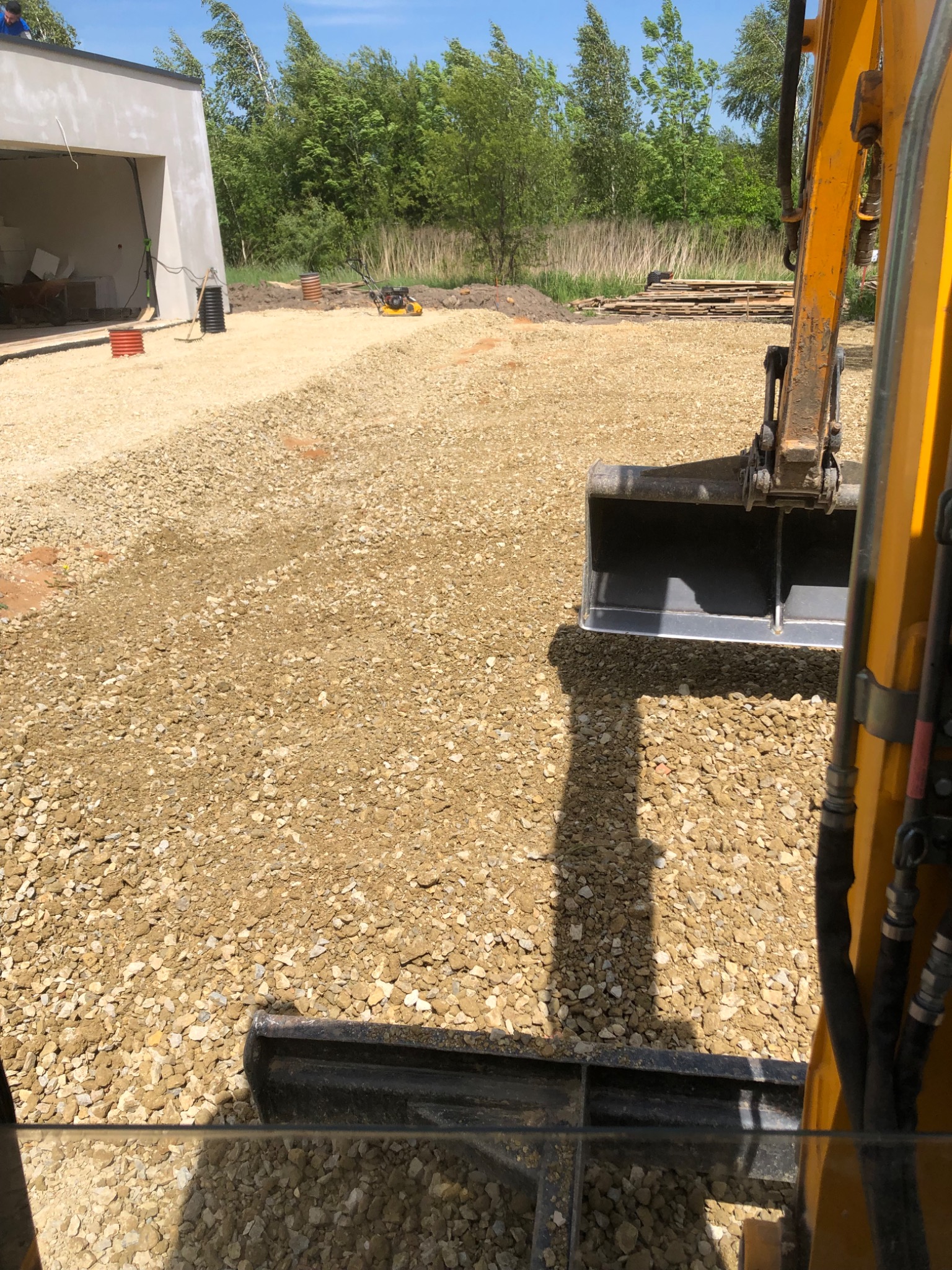 Widok z kabiny koparki na świeżo utwardzony teren żwirowy, w tle budynek w stanie surowym otwartym i zielone drzewa.