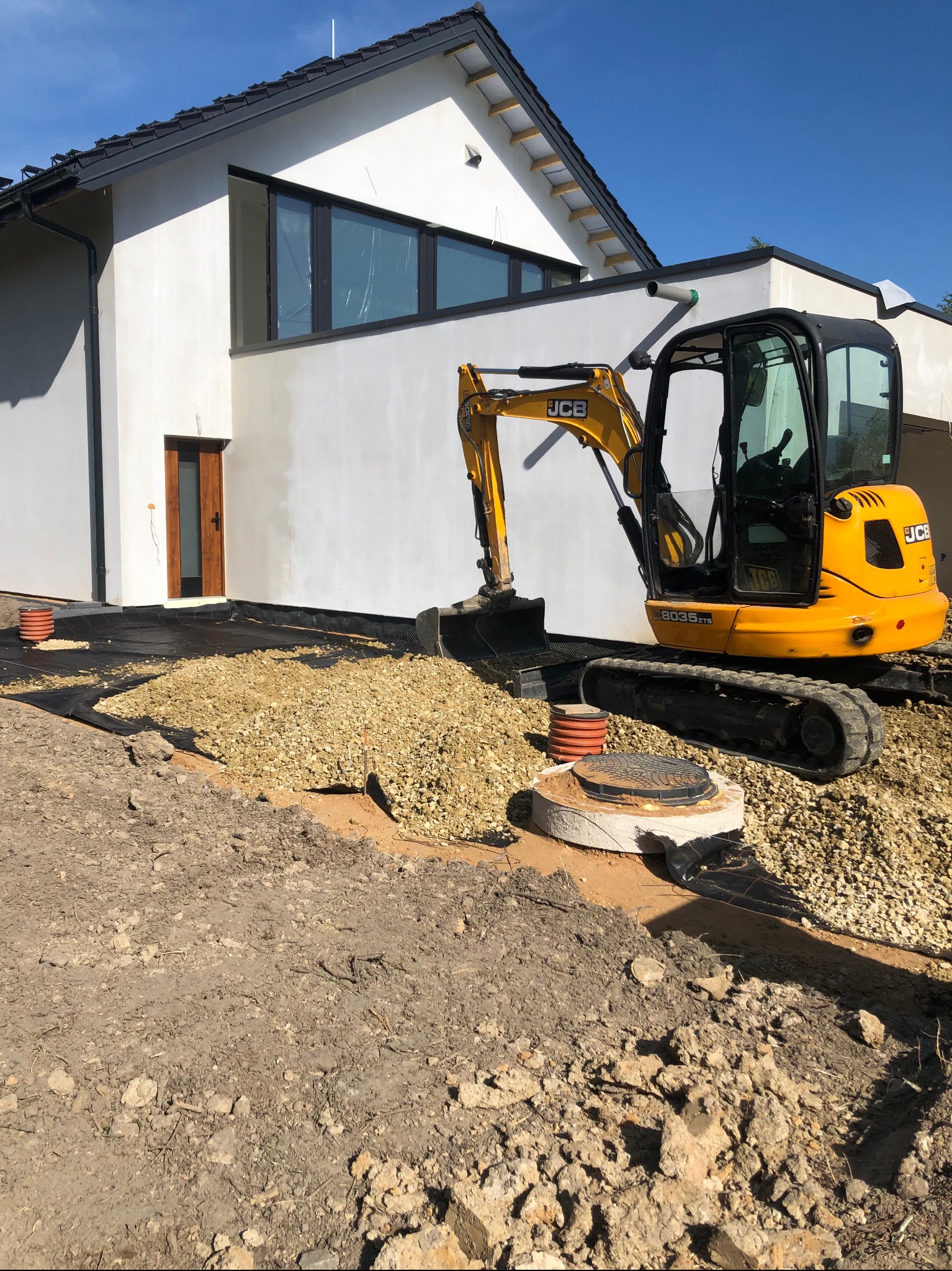 Żółta mini koparka JCB na tle nowoczesnego domu, przygotowująca teren pod instalacje, widoczny wykop, warstwa tłucznia i studzienka kanalizacyjna.
