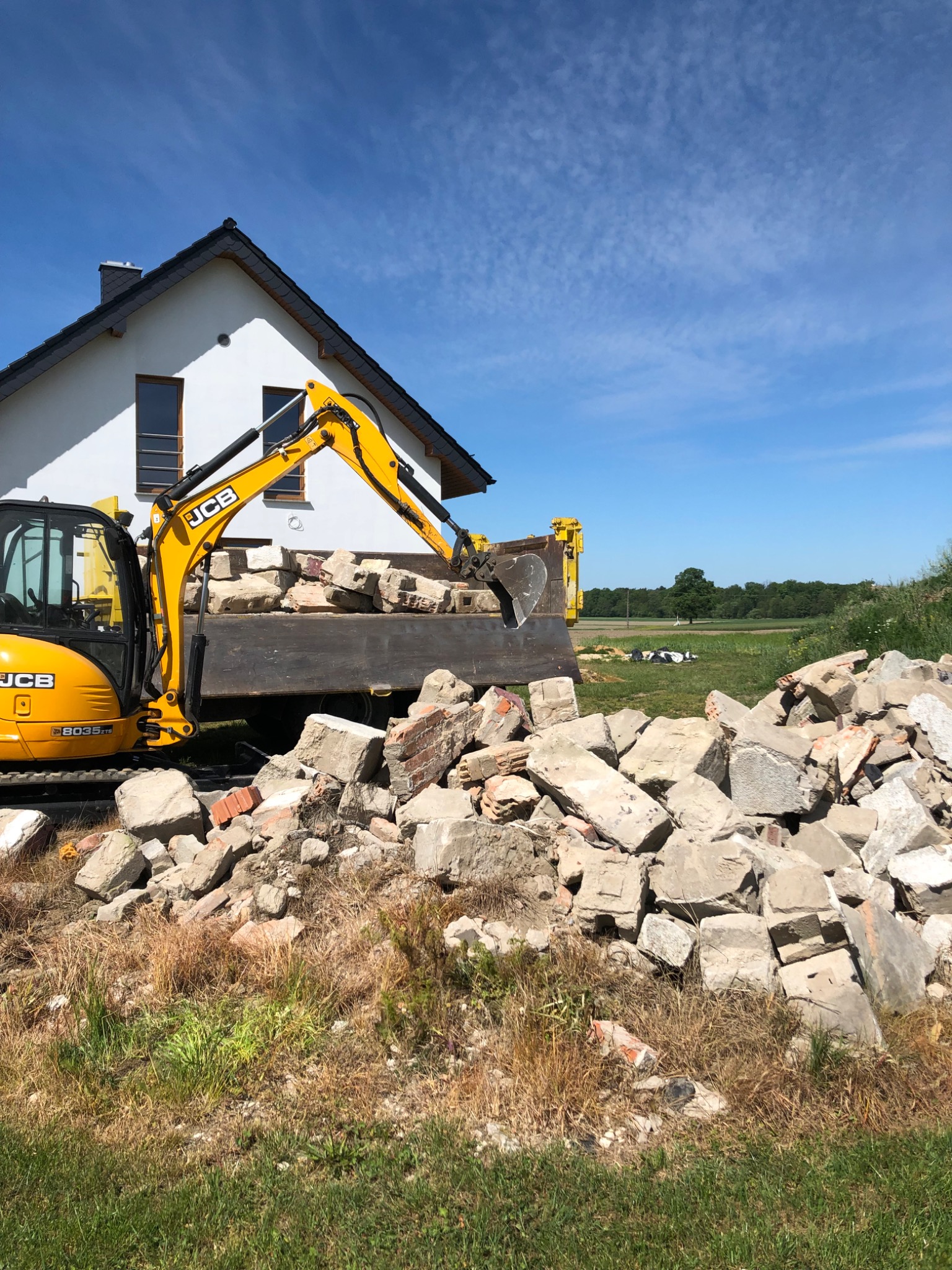 Żółta mini koparka JCB ładuje gruz betonowy i ceglany do skrzyni ciężarówki na tle białego domu z ciemnym dachem i błękitnego nieba.