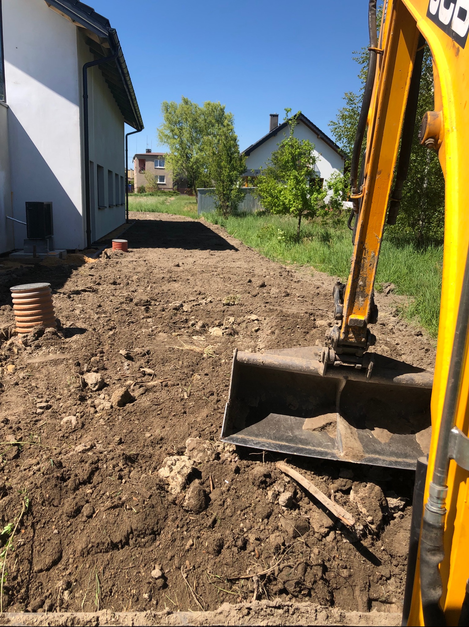 Żółta koparka z łyżką zagłębia się w ziemię na placu budowy, w tle budynek mieszkalny i niebieskie niebo. Widoczne pomarańczowe studzienki kanalizacyjne.