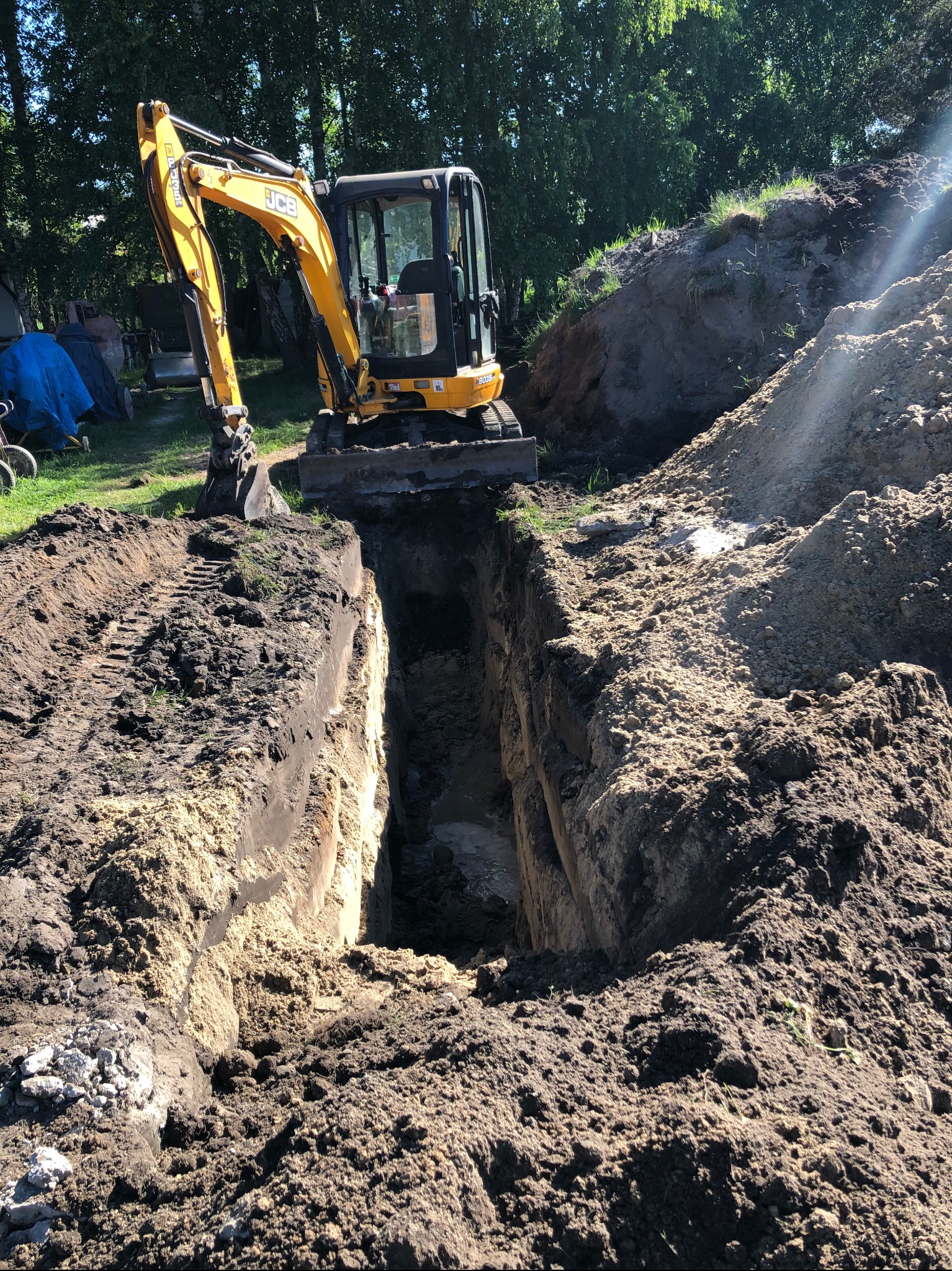 Żółta mini koparka JCB na tle zieleni drzew, wykonująca wąski głęboki wykop w ziemi, widoczne ślady opon i nasypy ziemi po obu stronach wykopu.