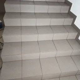 Schody wyłożone kwadratowymi, szarymi płytkami ceramicznymi z widocznymi fugami, prowadzące w górę wzdłuż białej ściany.