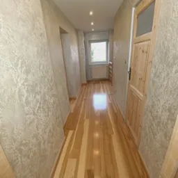 Korytarz po remoncie z drewnianą podłogą panelową i ścianami pokrytymi dekoracyjną, beżową strukturą. Widoczna drewniana ościeżnica i drzwi z szybą. W tle okno z grzejnikiem.