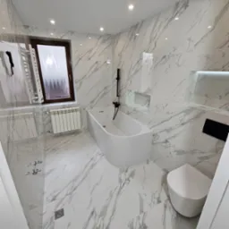 Nowoczesna łazienka wykończona białymi płytkami imitującymi marmur, z czarną armaturą i grzejnikiem drabinkowym, oświetlona punktowymi lampami sufitowymi.