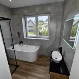 Nowoczesna łazienka z wanną wolnostojącą, szarą glazurą imitującą marmur i drewnopodobną podłogą. Okno z widokiem na wiejski krajobraz.