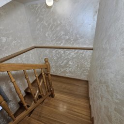 Usługi Remontowo-Budowlane Andrzej Knechta - Klatka schodowa z drewnianymi schodami i balustradą. Ściany wykończone dekoracyjnym tynkiem w odcieniach beżu i bieli. Na suficie widoczna lampa z trzema kulistymi kloszami.