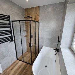 Usługi Remontowo-Budowlane Andrzej Knechta - Nowoczesna łazienka z kabiną prysznicową typu walk-in z czarnymi ramami, białą wanną i grzejnikiem drabinkowym na tle szarych płytek imitujących marmur.