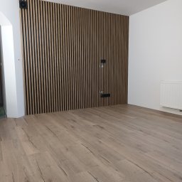 Usługi Remontowo-Budowlane Andrzej Knechta - Nowoczesne wykończenie wnętrza: drewniane lamele na ścianie z czarnymi akcentami, jasna podłoga z paneli, białe ściany i grzejnik. Minimalistyczny design.