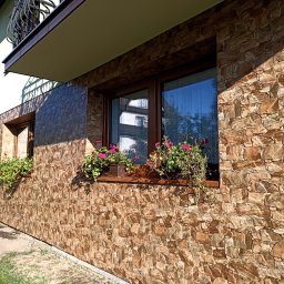 Usługi Remontowo-Budowlane Andrzej Knechta - Elewacja budynku wykończona kamieniem dekoracyjnym w odcieniach brązu i beżu. Okna z drewnianą ramą, ozdobione donicami z kwiatami. Widok z boku, w słoneczny dzień.