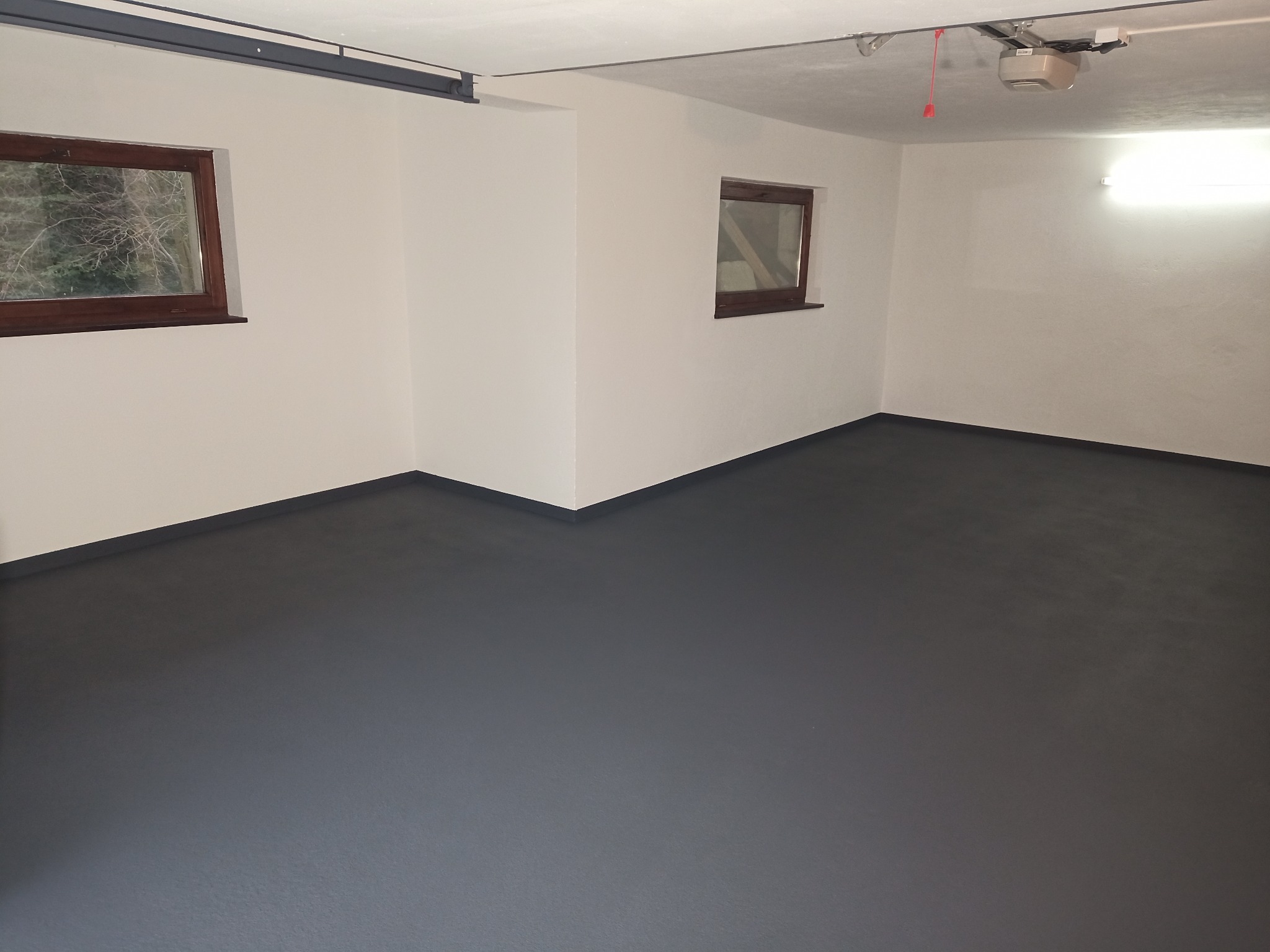 Wnętrze garażu po malowaniu ścian na biało i podłogi na szaro, z dwoma oknami w drewnianych ramach i zamontowanym napędem do bramy garażowej.